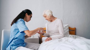 Gözlüklü esmer hemşire klinikteki yaşlı kadına hap veriyor.