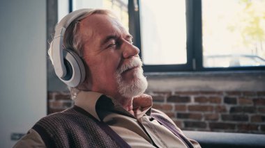 pleased senior man in wireless headphones listening music at home clipart