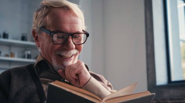 joyful senior man in eyeglasses reading book at home  clipart