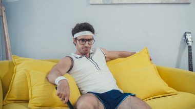 Spor giyim kuşağında tembel bir adam oturma odasında kanepede otururken televizyon izliyor.