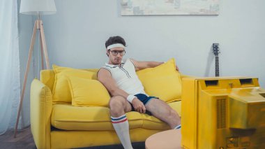 Spor giyim kuşağında tembel bir adam oturma odasındaki kanepede oturmuş retro TV izliyor.