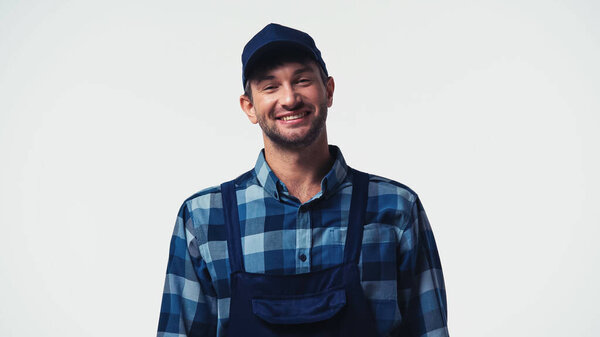 Cheerful handyman in overalls and cap looking at camera isolated on white