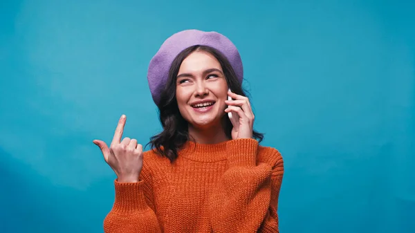 身着淡紫色贝雷帽的快乐女人一边对着自己指指点点 一边用蓝色的手机交谈 — 图库照片