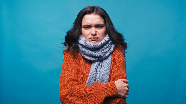 thick and frozen woman in warm scarf hugging herself isolated on blue