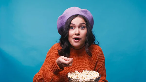 穿着套头毛衣和贝雷帽的女人看到令人兴奋的电影 吃着被蓝色隔离的爆米花 感到惊讶 — 图库照片
