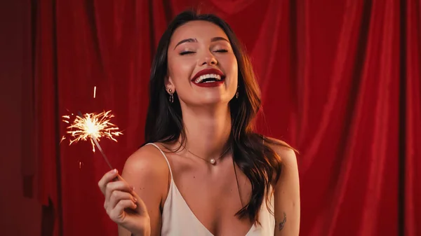 cheerful woman with tattoo holding bright sparkler and laughing on red
