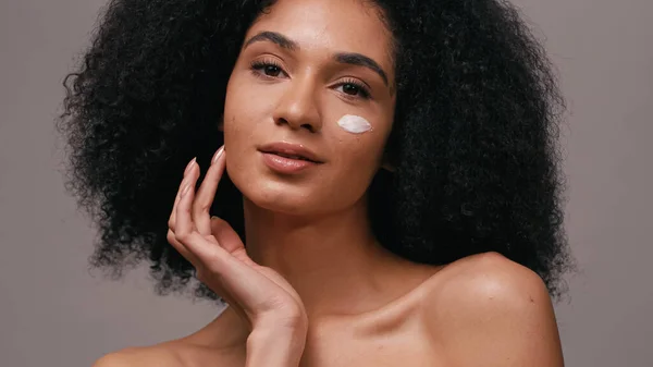 curly african american woman with cosmetic cream on face isolated on grey
