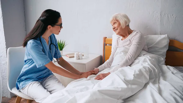 Geriatryczna Pielęgniarka Okularach Rozmawiająca Uśmiechniętą Starszą Kobietą Łóżku — Zdjęcie stockowe