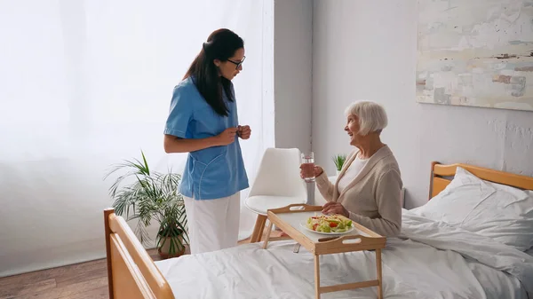 ブルネット看護師と話して幸せな高齢者近く朝食トレイ上のベッド — ストック写真