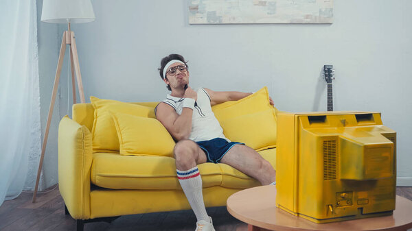 lazy man in sportswear scratching neck with remote controller while sitting on sofa in living room