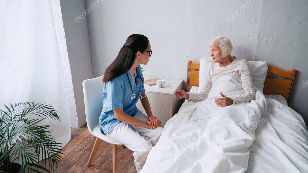 brunette nurse in eyeglasses and aged woman talking in clinic