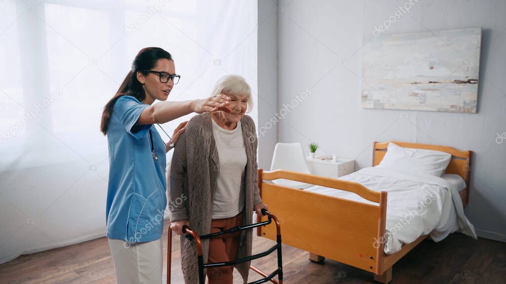 senior woman stepping with walkers near brunette nurse pointing away 