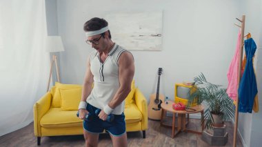 Spor gözlüklü ve spor giysili bir adam oturma odasında halterleri kaldırıyor.