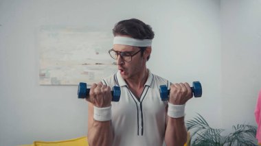 retro spor giyim ve gözlüklü adam evde dambıl kaldırıyor.