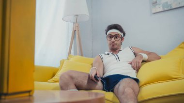 dissatisfied man in retro sportswear sitting on sofa and watching tv clipart