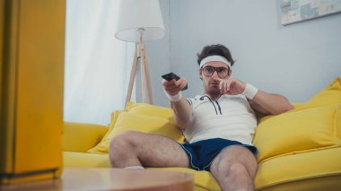 man in sportswear picking nose while sitting on sofa and clicking channels  clipart