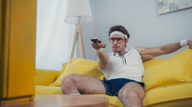 Gözlüklü ve retro spor giyimli bir adam kanepede oturuyor ve kanalları değiştiriyor. 