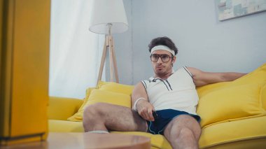 Eski spor kıyafetleri içinde sıkılmış bir adam kanepede oturuyor ve kanal değiştiriyor. 