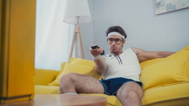 Eski spor kıyafetleri içinde sıkılmış bir adam kanepede oturuyor ve kanal değiştiriyor. 