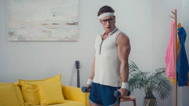 Şok olmuş sporcu oturma odasında dambıllarla çalışıyor, retro spor konsepti.