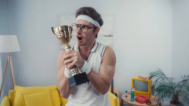Şok olmuş gözlüklü sporcu evinde altın kupa tutuyor.