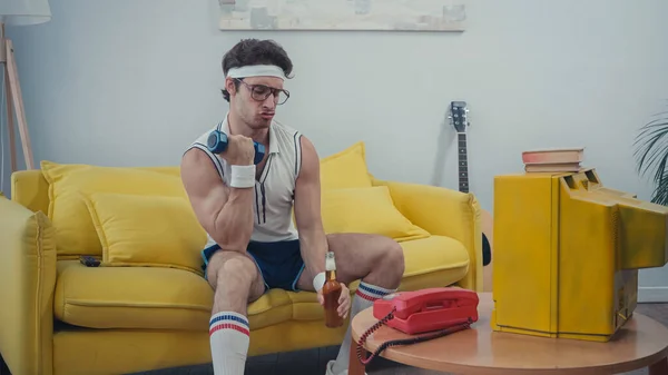 sportive man drinking beer and lifting dumbbell while sitting on sofa