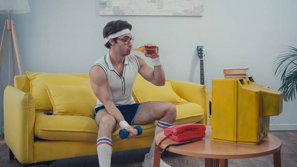 sportsman drinking beer and exercising with dumbbell while watching tv