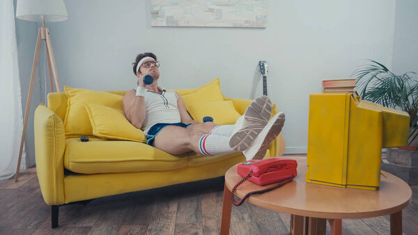 Sportsman lifting dumbbells while sitting on couch near retro tv in living room