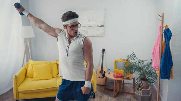 Sportsman in eyeglasses and retro sportswear lifting dumbbells in living room