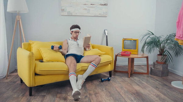 sportive man lifting dumbbell while reading book in living room