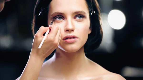 Makeup artist applying eye shadow on lower eyelid of model with bare shoulders — Stock Photo