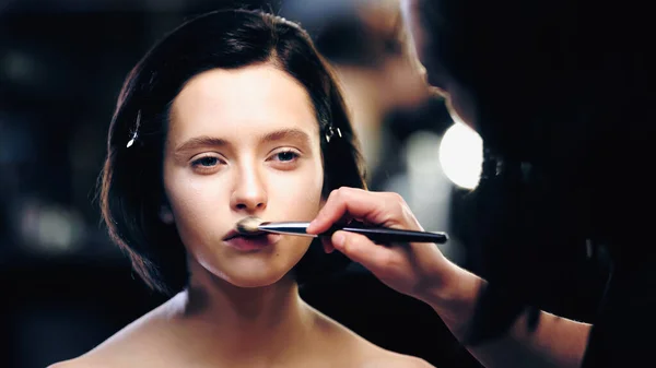 Artista de maquiagem aplicando cosméticos decorativos no rosto de modelo bonito com ombros nus — Fotografia de Stock