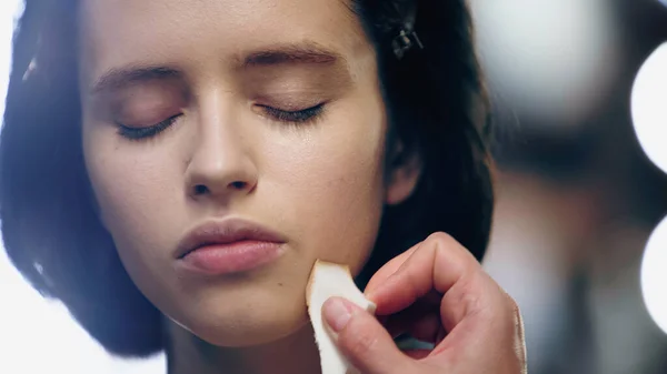 Maquillaje artista aplicando maquillaje fundación con esponja cosmética en la cara de la mujer con los ojos cerrados - foto de stock