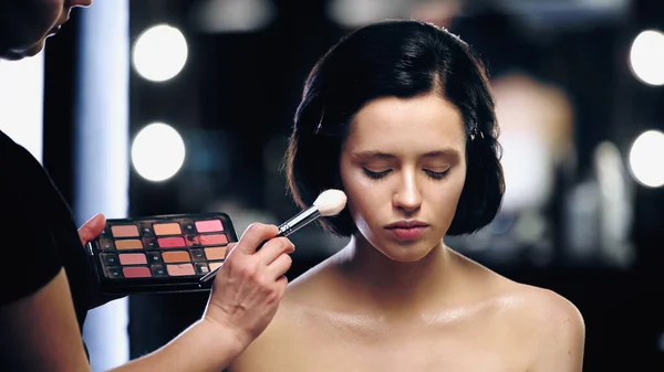 Artista de maquiagem segurando paleta e aplicando iluminador na pele do modelo com escova cosmética — Fotografia de Stock