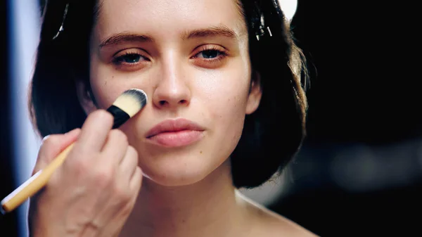 Maquilleur tenant pinceau cosmétique et appliquant fond de teint de maquillage sur le visage de la femme — Photo de stock