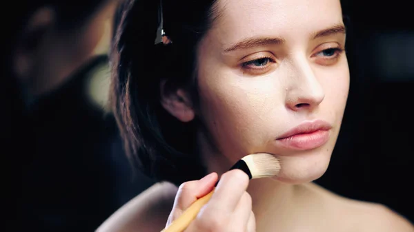 Trucco sfocato artista tenendo pennello cosmetico mentre si applica fondazione trucco sulla pelle del giovane modello — Foto stock