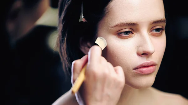 Blurred makeup artist holding cosmetic brush while applying makeup foundation on face of young brunette model — Stock Photo