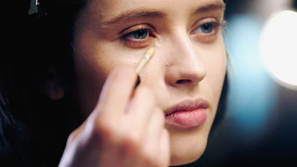 Makeup artist holding cosmetic brush while applying concealer on skin of young brunette model — Stock Photo