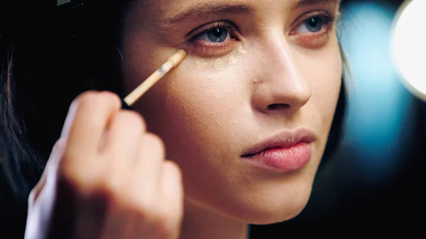 Borrosa mano de maquillador sosteniendo cepillo cosmético mientras se aplica corrector en la cara de la joven modelo morena - foto de stock