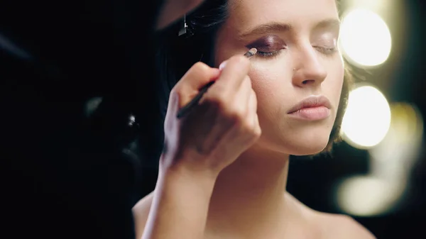 Blurred makeup artist applying dark eye shadow with cosmetic brush on eyelids of model with closed eyes — Stock Photo