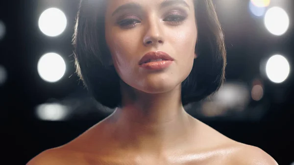 Bonita mujer joven con maquillaje y hombros desnudos mirando a la cámara con luz de fondo - foto de stock