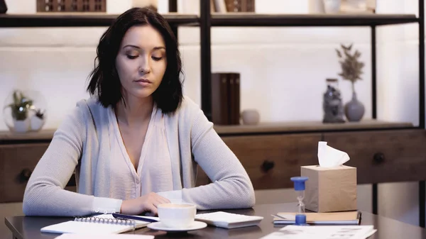 Thoughtful woman sitting on table with cup of coffee, calculator, sandglass and notebooks with pen at home — Stock Photo