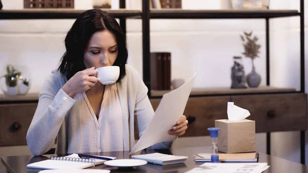 Morena sentada en la mesa, bebiendo café y mirando documentos en casa - foto de stock
