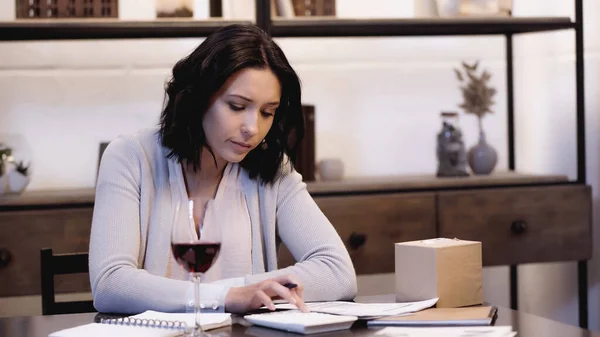 Upset woman sitting on table with glass of red wine and counting on calculator at home — Stock Photo