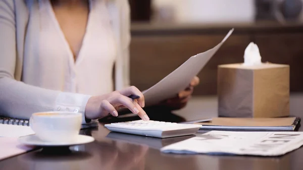 Partial view of woman sitting on table with documents and counting on calculator with finger at home — Stock Photo