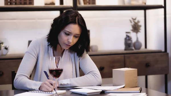 Serious woman sitting on table with glass of red wine and writing in notebook at home — Stock Photo