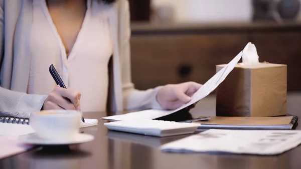 Partial view of woman sitting on table, holding documents and writing with pen in notebook at home — Stock Photo