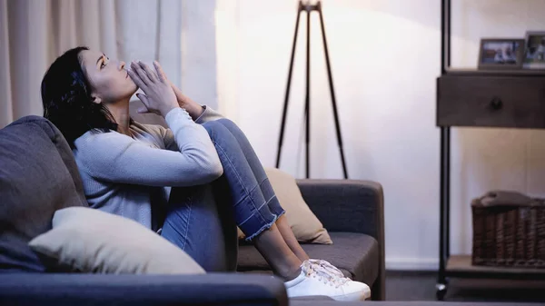 Depressed woman in casual clothes sitting on sofa with praying hands near mouth at home — Stock Photo