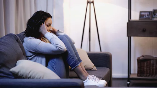 Mulher deprimida em roupas casuais sentado no sofá e de mãos dadas perto da cabeça em casa — Fotografia de Stock