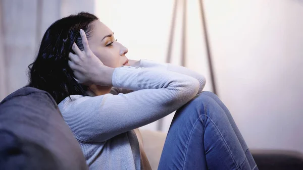 Mujer deprimida en ropa casual sentada en el sofá y cogida de la mano cerca de la cabeza en casa - foto de stock